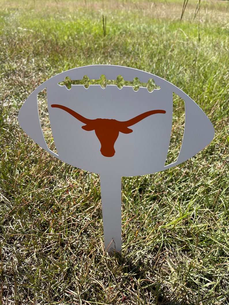 Peut inclure: Un piquet de jardin en m&eacute;tal blanc en forme de ballon de football, orn&eacute; d'un logo de longhorn orange br&ucirc;l&eacute;. Le piquet pr&eacute;sente des d&eacute;tails d&eacute;coup&eacute;s et est plant&eacute; dans l'herbe, id&eacute;al pour la d&eacute;coration ext&eacute;rieure.