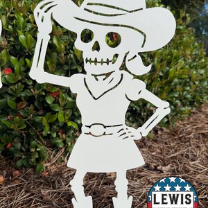 May include: White metal cutout of a skeleton in a cowboy hat, dress, and boots. The skeleton is posed with one arm raised to the hat and the other on its hip. The cutout is placed in a garden setting. A circular logo with the text "LEWIS DESIGN COMPANY" is in the bottom right.