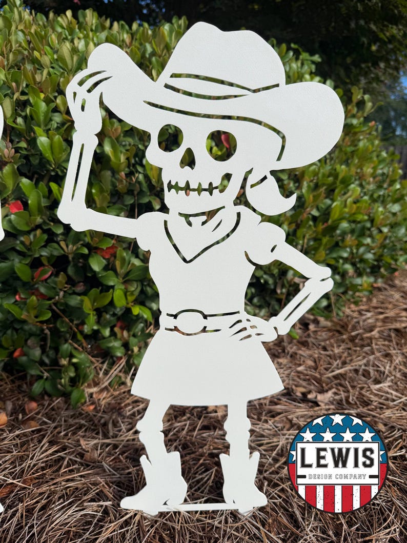 May include: White metal cutout of a skeleton in a cowboy hat, dress, and boots. The skeleton is posed with one arm raised to the hat and the other on its hip. The cutout is placed in a garden setting. A circular logo with the text "LEWIS DESIGN COMPANY" is in the bottom right.