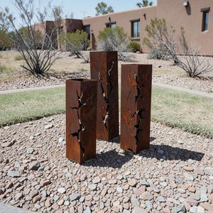 May include: Three weathered, rectangular metal sculptures with a rusted finish. Each sculpture features a vertical, crack-like design. The sculptures are placed outdoors on a bed of small rocks, with a grassy area and a building in the background.