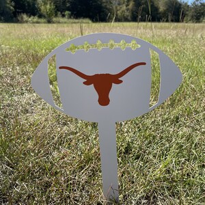Peut inclure: Un piquet de jardin en m&eacute;tal blanc en forme de ballon de football avec un logo de longhorn orange br&ucirc;l&eacute;. Le piquet est plant&eacute; dans un champ herbeux sous un ciel bleu clair. Le ballon de football a des d&eacute;tails de couture verts.
