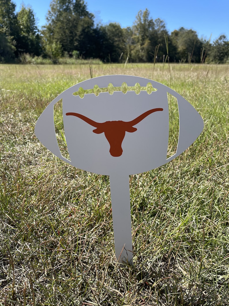 Peut inclure: Un piquet de jardin en m&eacute;tal blanc en forme de ballon de football avec un logo de longhorn orange br&ucirc;l&eacute;. Le piquet est plant&eacute; dans un champ herbeux sous un ciel bleu clair. Le ballon de football a des d&eacute;tails de couture verts.