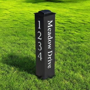 May include: A black solar-powered address sign with the numbers 1234 and the words "Meadow Drive" in white. The sign is rectangular and stands upright on a green lawn.