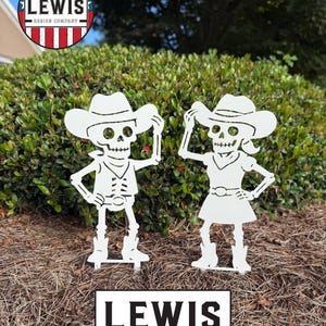 May include: Two white metal cowboy skeleton garden ornaments. One skeleton wears a vest and the other a dress. The ornaments are in front of a green bush. The text "LEWIS DESIGN COMPANY" is at the bottom.
