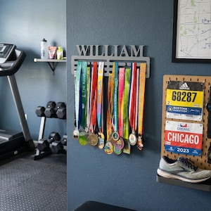 May include: A home gym with a treadmill, dumbbells, and a medal display. The medal holder has the name "WILLIAM" above a row of colorful race medals. Race bibs and running shoes are also displayed.
