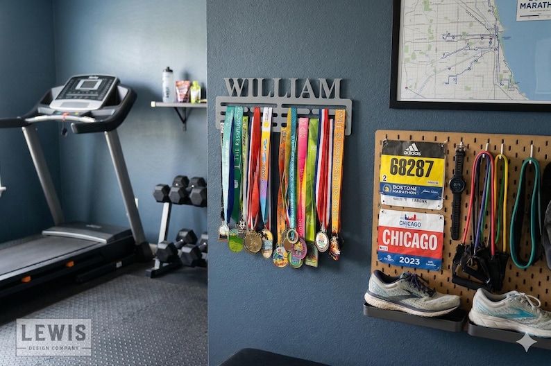 May include: A home gym with a treadmill, dumbbells, and a medal display. The medal holder has the name "WILLIAM" above a row of colorful race medals. Race bibs and running shoes are also displayed.