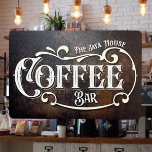 May include: A vintage-style metal sign with a brown background and white lettering. The sign reads "The Java House Coffee Bar" with decorative swirls and flourishes.