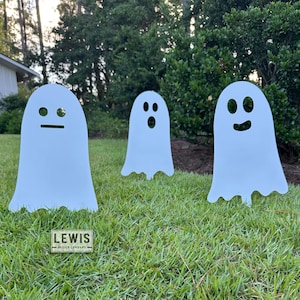 May include: Three white ghost yard decorations with different facial expressions are displayed on a green lawn. The ghosts are cut from a flat material and have a simple, cartoonish design. A small sign with the text "LEWIS DESIGN COMPANY" is at the bottom.