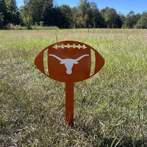 Peut inclure: Un piquet de jardin en m&eacute;tal orange en forme de ballon de football, orn&eacute; d'un logo de longhorn blanc. Le piquet est plant&eacute; dans un champ herbeux sous un ciel bleu. Le design est d&eacute;coup&eacute; pour ressembler aux lacets d'un ballon.