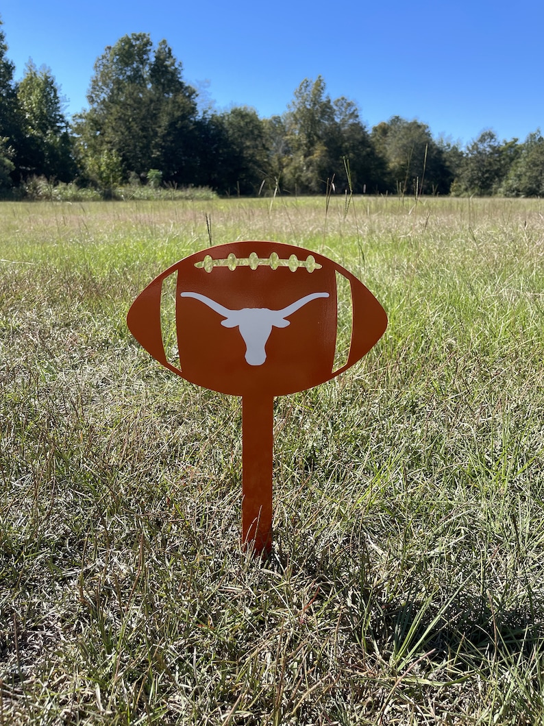 Peut inclure: Un piquet de jardin en m&eacute;tal orange en forme de ballon de football, orn&eacute; d'un logo de longhorn blanc. Le piquet est plant&eacute; dans un champ herbeux sous un ciel bleu. Le design est d&eacute;coup&eacute; pour ressembler aux lacets d'un ballon.