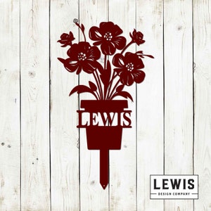 May include: A red metal flower pot with the name "LEWIS" in white letters. The pot is decorated with a bouquet of red flowers and green leaves. The pot is attached to a metal stake for planting in the ground.