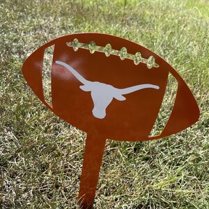 Peut inclure: Un piquet de jardin en m&eacute;tal orange en forme de ballon de football avec un logo de longhorn blanc. Le ballon de football pr&eacute;sente des d&eacute;tails d&eacute;coup&eacute;s et est plac&eacute; dans une zone herbeuse. Un objet d&eacute;coratif pour les fans de sport.
