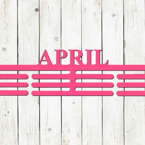 May include: A bright pink metal sign with the word "APRIL" at the top. Below the word are three horizontal bars with evenly spaced slots, designed to hold items. The sign is mounted on a white wooden plank background.