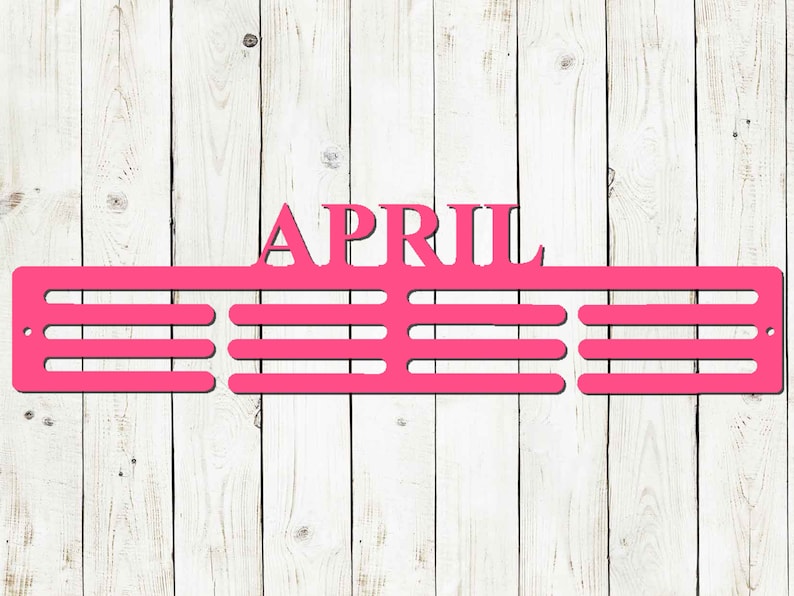 May include: A bright pink metal sign with the word "APRIL" at the top. Below the word are three horizontal bars with evenly spaced slots, designed to hold items. The sign is mounted on a white wooden plank background.