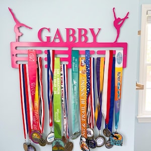 May include: A pink metal medal display with the name "GABBY" and gymnast silhouettes. The display holds numerous medals with colorful ribbons, showcasing achievements from various races and events. The display is mounted on a light blue wall.