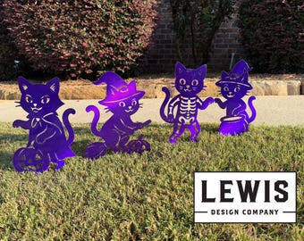 May include: Four purple metal Halloween cat yard decorations. One cat holds a pumpkin, another rides a broom, one is a skeleton, and the last stirs a cauldron. The Lewis Design Company logo is in the lower right.