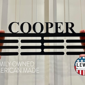 May include: Black metal sign with the name "COOPER" and three horizontal bars. The sign is held up by hands. The text "FAMILY OWNED AMERICAN MADE" is below the sign, and a circular logo is in the bottom right corner.