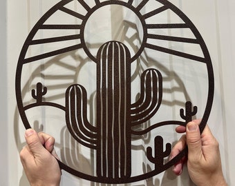 Puede incluir: Una pieza de arte de pared de metal con un cactus estilizado con un sol y montañas en el fondo. El arte es de forma circular y de color marrón. El texto "LEWIS DESIGN COMPANY" está en la parte superior de la imagen. El texto "FAMILY OWNED AMERICAN MADE" está en la parte inferior de la imagen.