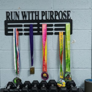 May include: Black metal medal display with the words "RUN WITH PURPOSE" above a series of slots holding colorful race medals. The medals have various colored ribbons and designs. The display is mounted on a gray brick wall.