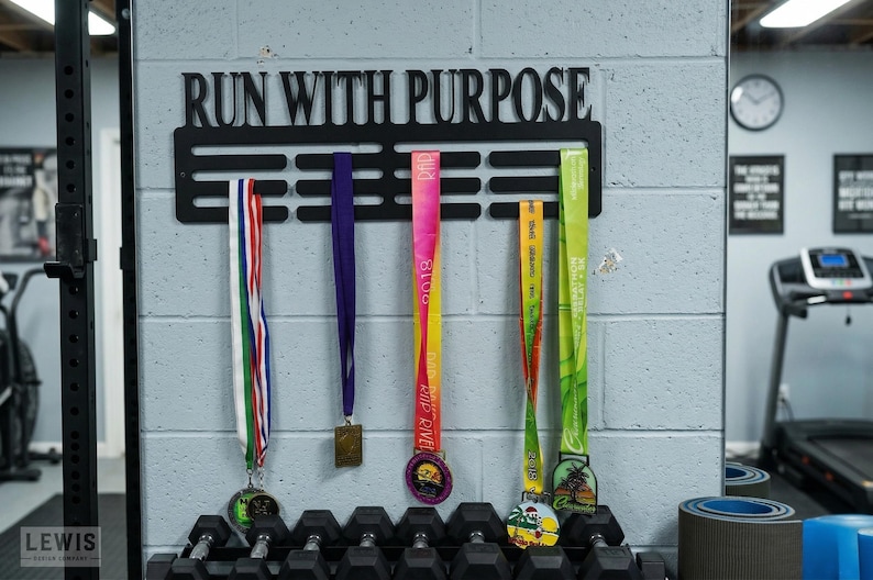 May include: Black metal medal display with the words "RUN WITH PURPOSE" above a series of slots holding colorful race medals. The medals have various colored ribbons and designs. The display is mounted on a gray brick wall.