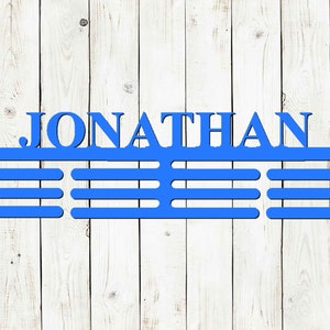May include: A blue metal medal display with the name "JONATHAN" cut out at the top. Below the name are three horizontal bars for hanging medals. The display is mounted on a white wooden background.