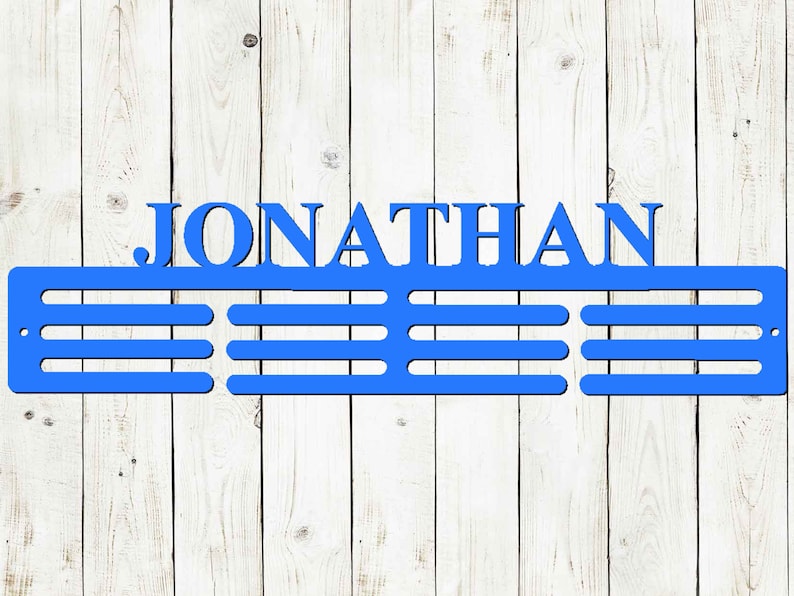 May include: A blue metal medal display with the name "JONATHAN" cut out at the top. Below the name are three horizontal bars for hanging medals. The display is mounted on a white wooden background.