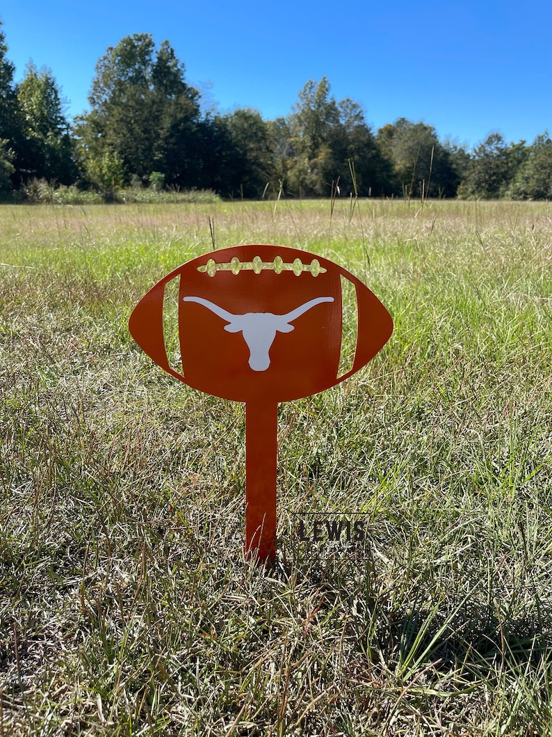 Peut inclure: Un piquet de jardin en m&eacute;tal orange en forme de ballon de football avec un logo de longhorn blanc. Le piquet est plant&eacute; dans un champ herbeux sous un ciel bleu clair. Le ballon a un design d&eacute;coup&eacute;.