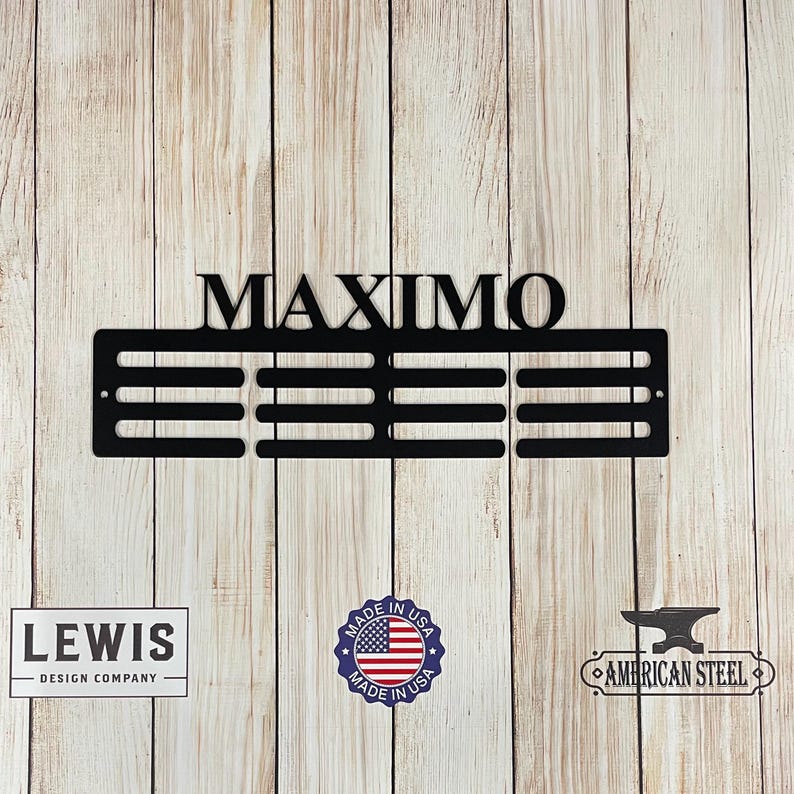 May include: Black metal wall decor with the name "MAXIMO" above a series of horizontal bars. The design includes logos for "LEWIS DESIGN COMPANY", "MADE IN USA", and "AMERICAN STEEL". The piece is mounted on a light wood background.