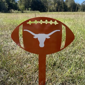Peut inclure: Un piquet de jardin en m&eacute;tal orange en forme de ballon de football avec un logo de longhorn blanc. Le design du ballon de football pr&eacute;sente des d&eacute;tails d&eacute;coup&eacute;s et est plant&eacute; dans un champ herbeux sous un ciel bleu.