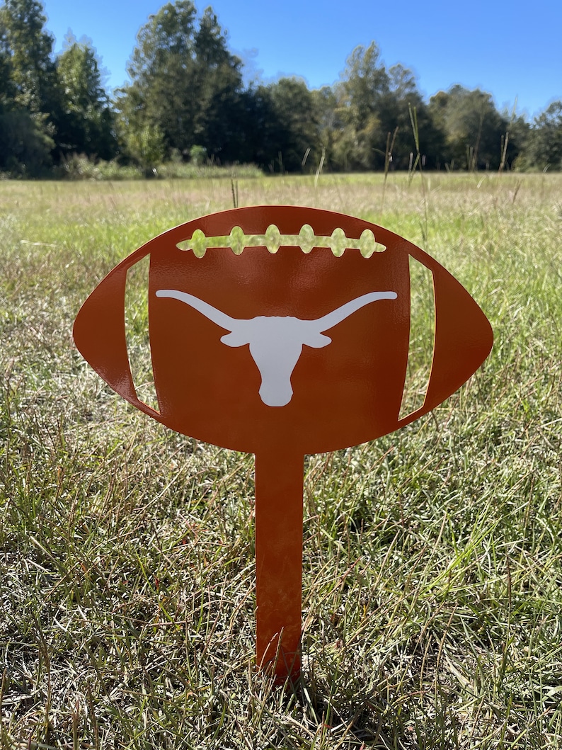 Peut inclure: Un piquet de jardin en m&eacute;tal orange en forme de ballon de football avec un logo de longhorn blanc. Le design du ballon de football pr&eacute;sente des d&eacute;tails d&eacute;coup&eacute;s et est plant&eacute; dans un champ herbeux sous un ciel bleu.