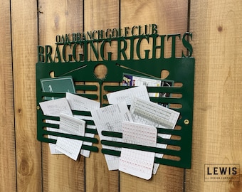 May include: Green metal golf scorecard holder with the text "Oak Branch Golf Club Bragging Rights". The holder has slots for holding scorecards and is mounted on a wood wall.