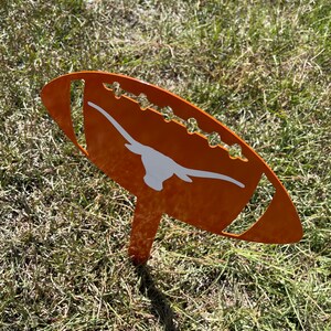 Peut inclure: Un piquet de jardin en m&eacute;tal orange en forme de ballon de football, orn&eacute; d'un logo de longhorn blanc. Le ballon de football a des coutures d&eacute;coratives et est plant&eacute; dans l'herbe.