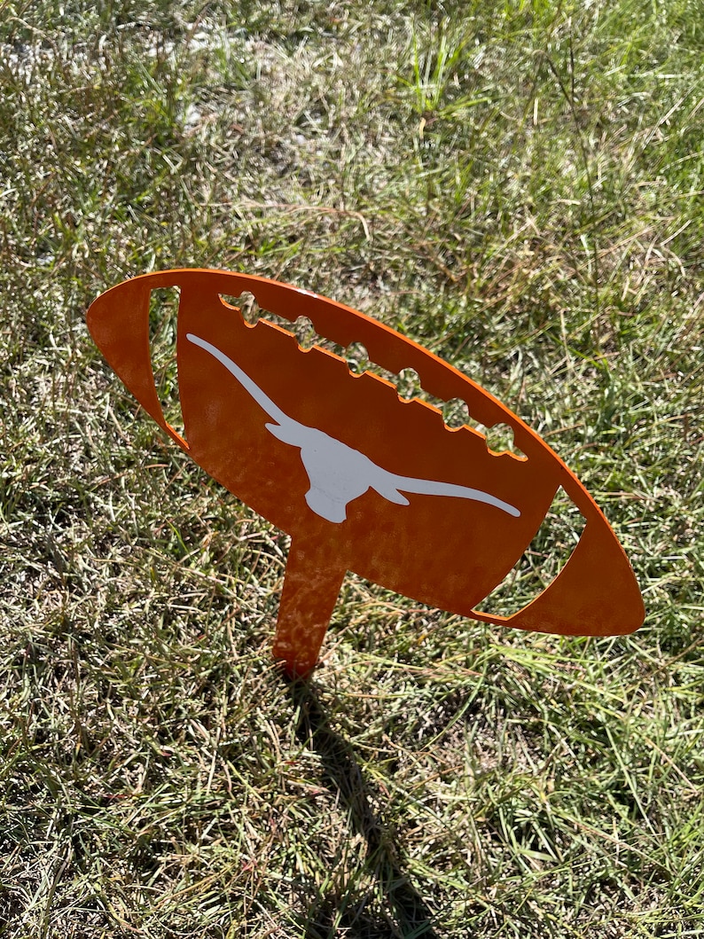 Peut inclure: Un piquet de jardin en m&eacute;tal orange en forme de ballon de football, orn&eacute; d'un logo de longhorn blanc. Le ballon de football a des coutures d&eacute;coratives et est plant&eacute; dans l'herbe.