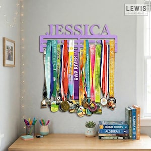 May include: A lavender-colored medal display with the name "JESSICA" cut out at the top. Colorful ribbons and medals hang from the display. The display is mounted on a gray wall above a wooden desk.