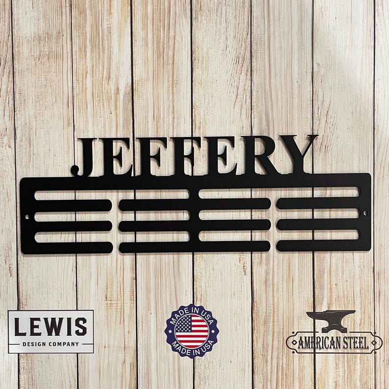 May include: Black metal wall art with the name "JEFFERY" cut out at the top, with three horizontal rows of slots below. The piece is mounted on a wooden background. Logos for "LEWIS DESIGN COMPANY", "MADE IN USA", and "AMERICAN STEEL" are visible.