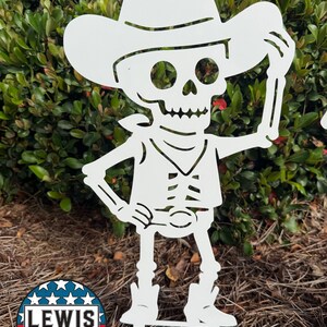 May include: White metal garden art of a skeleton cowboy wearing a hat and boots. The skeleton is holding up the brim of its hat. The design includes a vest, a neckerchief, and a belt buckle. The Lewis Design Company logo is in the lower left corner.