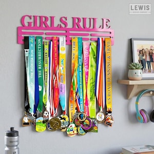May include: A pink metal medal display with the words "GIRLS RULE" across the top. The display holds numerous colorful race medals with ribbons. A water bottle and framed artwork are visible in the background.