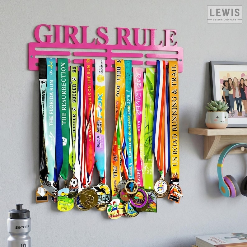May include: A pink metal medal display with the words "GIRLS RULE" across the top. The display holds numerous colorful race medals with ribbons. A water bottle and framed artwork are visible in the background.