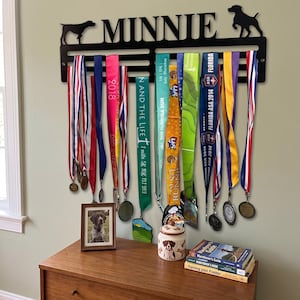 May include: A black metal wall-mounted medal hanger featuring the name MINNIE in capital letters, flanked by two dog silhouettes. Multiple colorful race medals with ribbons hang from the rack. Below sits a framed dog photo, a ceramic jar, and books.