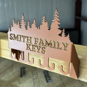 May include: A metal key holder with a forest silhouette design. The holder has the text "Smith Family Keys" in gold lettering and five hooks for hanging keys.