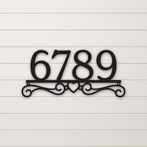 May include: Black metal house number plaque with the numbers 6789 and a heart design. The plaque is mounted on a white wooden wall.