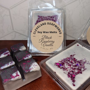May include: Soy wax melts in a clear plastic container. The wax melts are black and white with pink glitter and are labeled "Cleveland Soapworks - Soy Wax Melts - Black Raspberry Vanilla - 2.75 FL. OZ."