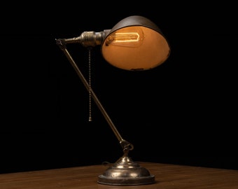 May include: An antique-style desk lamp with a brass finish and a dark, curved shade. The lamp features an exposed Edison-style bulb, a pull chain, and adjustable arm. The lamp is on a wooden surface against a black background.
