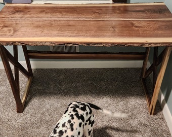 May include: A live edge walnut desk with a natural wood finish and a simple X-shaped base. The desk is made of solid wood and has a rustic look. The desk is sitting on a carpeted floor.