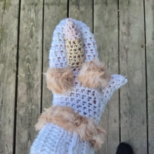 May include: A white crochet mitten featuring a central gold-toned horn, two fluffy tan faux fur pom-poms, and a matching tan faux fur cuff. The mitten is constructed with a textured stitch pattern and includes a thumb section on the right side.