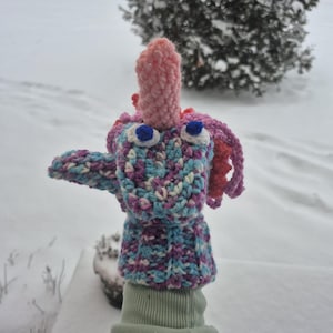 May include: A handmade crochet monster puppet in shades of blue, purple, and pink. The puppet has large blue eyes, a pink nose, and purple hair. The puppet is being held up in front of a snowy background.