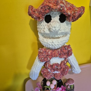 May include: A crocheted puppet with a white head and body, featuring large black eyes and a prominent nose. It wears a ruffled hat and a matching bow tie in a variegated yarn of pink, orange, and brown. The puppet is positioned above small decorative mushroom figures and artificial pink flowers.
