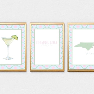 May include: Three framed art prints with gold frames. The prints feature a cocktail with a lime, the text "Chapel Hill, North Carolina", and a map of North Carolina with the text "Go To Heck". The frames have a pink and blue patterned border.