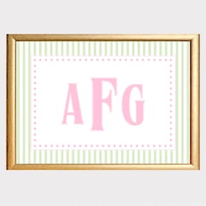 May include: A rectangular print in a gold frame featuring the letters AFG in large pink serif font. The center is white with a pink dotted border, surrounded by a background of vertical light green and white stripes on the print itself.