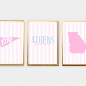 May include: Three framed prints with gold borders. The prints feature a pink and white striped background. One print has a pennant with "1785", another says "Athens Georgia", and the last is a pink Georgia state outline.